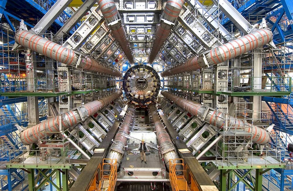 Detector ATLAS em construção: um enorme cilindro com oito tubos em volta, com uma pessoa pequena embaixo para comparar