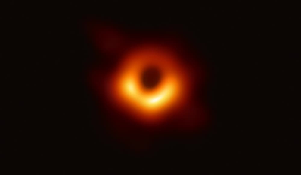 Anel laranja brilhante com o centro escuro, sobre fundo preto: a imagem do buraco negro M87