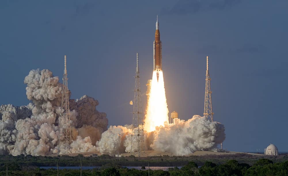 O foguete SLS subindo com chamas fortes e muita fumaça branca, entre torres na base de lançamento