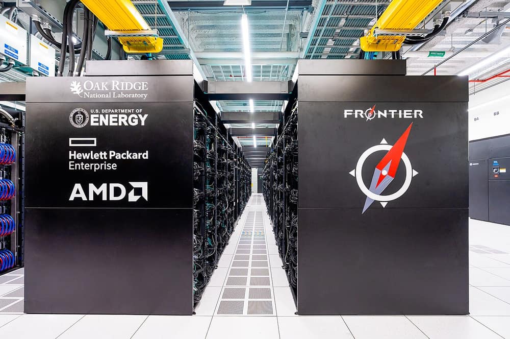 Corredor entre armários pretos do supercomputador Frontier, com os logotipos do laboratório de Oak Ridge