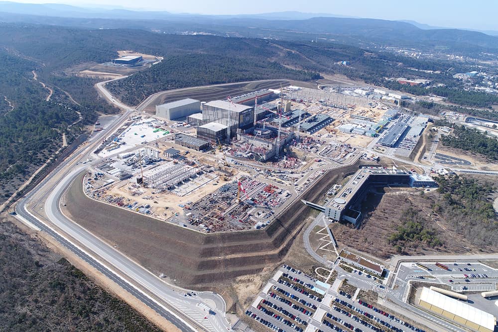 Vista aérea de um grande canteiro de obras com prédios industriais e guindastes, cercado de mata