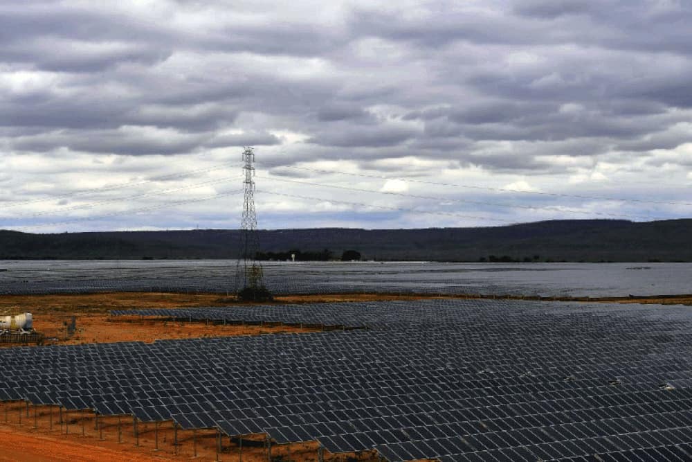 Milhares de painéis solares num terreno plano, com uma torre de transmissão e o céu nublado