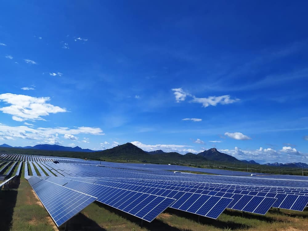 Fileiras de painéis solares azuis sob um céu azul com nuvens e morros ao fundo
