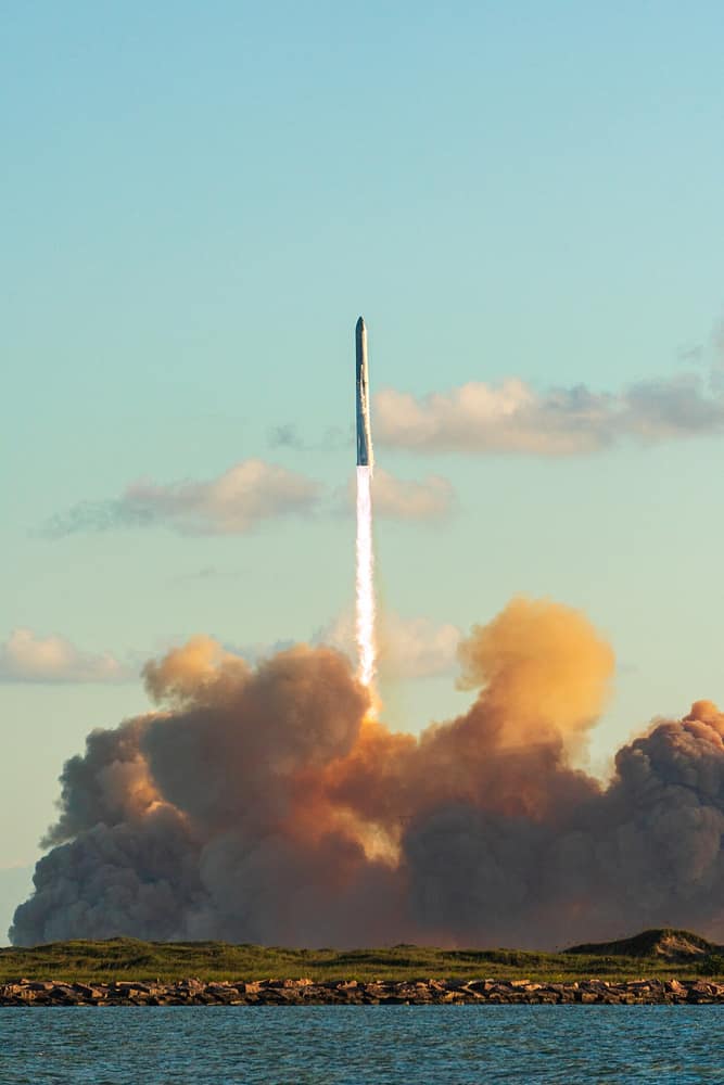 O foguete Starship subindo no céu azul, com uma nuvem enorme de fumaça alaranjada embaixo