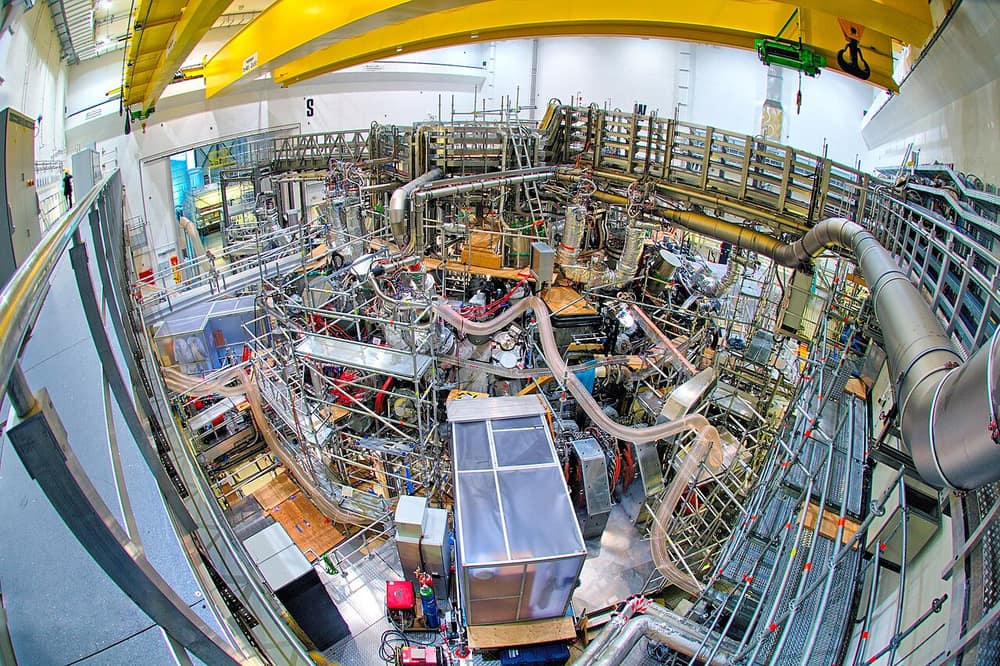 Máquina gigante cheia de tubos, cabos e plataformas dentro de um salão, vista de cima