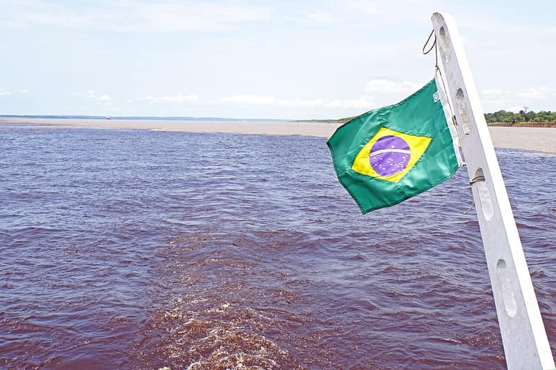 Rio de água escura encontrando outro de água barrenta, com a bandeira do Brasil num barco