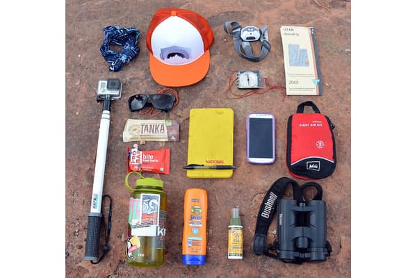 Itens de trilha arrumados no chão: boné, óculos escuros, lanche, garrafa de água, protetor solar, celular, mapa, bússola, kit de primeiros socorros e binóculo