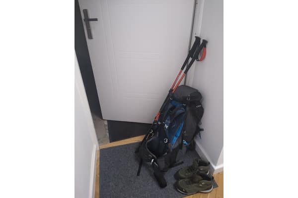 Mochila de trilha encostada na parede, com bastões de caminhada e botas no chão